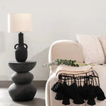 Black ceramic table lamp on a stacked stone side table beside a beige armchair with a tasseled throw- Summer sale Dubai