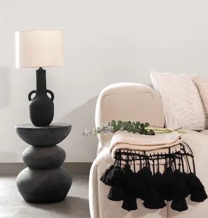 Black ceramic table lamp on a stacked stone side table beside a beige armchair with a tasseled throw- Summer sale Dubai