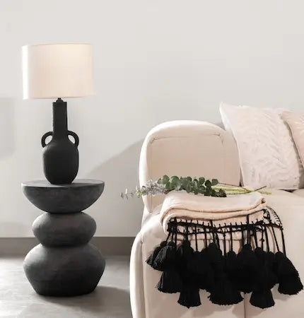 Black ceramic table lamp on a stacked stone side table beside a beige armchair with a tasseled throw- Summer sale Dubai